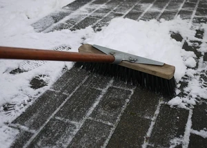 Ein Besen fegt Schnee von einem gepflasterten Gehweg, auf dem noch Fußspuren im Schnee zu sehen sind.