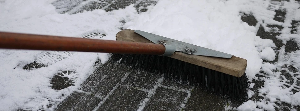 Ein Besen fegt Schnee von einem gepflasterten Gehweg, auf dem noch Fußspuren im Schnee zu sehen sind.