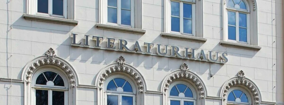 Fassade des Literaturhauses Hamburg Das Bild zeigt die Fassade eines klassizistisch anmutenden Gebäudes mit verzierten, rundbogigen Fenstern. An der Fassade ist in großen, metallenen Buchstaben das Wort „LITERATURHAUS“ angebracht. Vor dem unteren Teil der Fassade befindet sich ein schmiedeeiserner Balkon mit Ziergittern.