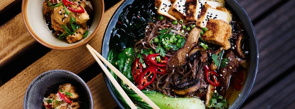 Eine Schale mit asiatischer Nudelsuppe, garniert mit gebratenem Tofu, Pak Choi, Pilzen, frischen Kräutern, Frühlingszwiebeln, Chilischeiben und Sesam. Daneben stehen zwei kleine Schälchen mit frittierten Tofu-Stücken, ebenfalls mit Sesam, Kräutern und Chili dekoriert. Ein Paar Essstäbchen liegt auf der großen Schale.