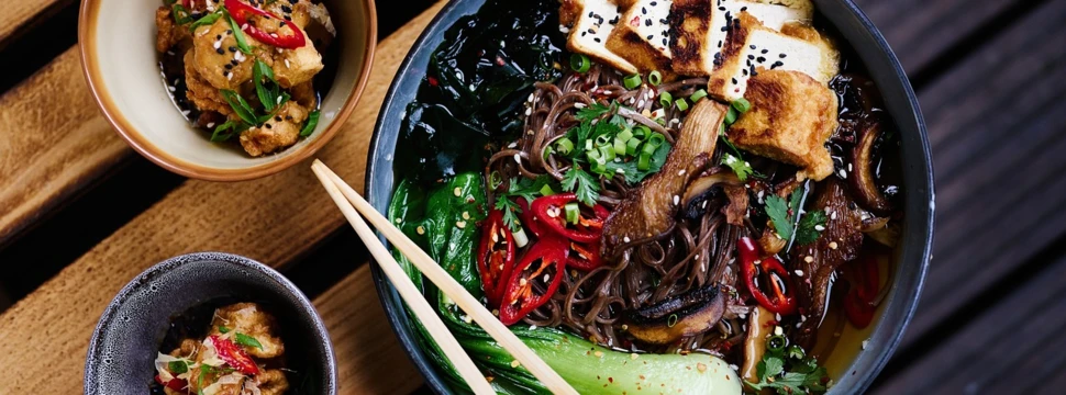 Eine Schale mit asiatischer Nudelsuppe, garniert mit gebratenem Tofu, Pak Choi, Pilzen, frischen Kräutern, Frühlingszwiebeln, Chilischeiben und Sesam. Daneben stehen zwei kleine Schälchen mit frittierten Tofu-Stücken, ebenfalls mit Sesam, Kräutern und Chili dekoriert. Ein Paar Essstäbchen liegt auf der großen Schale.