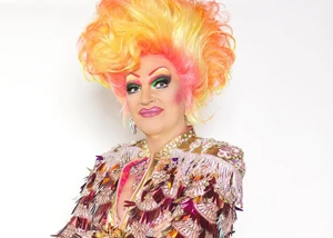 Olivia Jones mit gelb‑orangefarbener Hochfrisur, auffälligem Make-up und glitzerndem, mehrfarbigem Paillettenkostüm vor hellem Hintergrund.