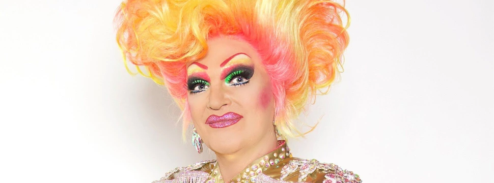 Olivia Jones mit gelb‑orangefarbener Hochfrisur, auffälligem Make-up und glitzerndem, mehrfarbigem Paillettenkostüm vor hellem Hintergrund.