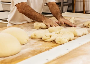 Hände eines Bäckers formen Teiglinge auf einer bemehlten Arbeitsfläche in einer Backstube. Im Hintergrund sind Arbeitsutensilien und Regale mit Behältern zu sehen.