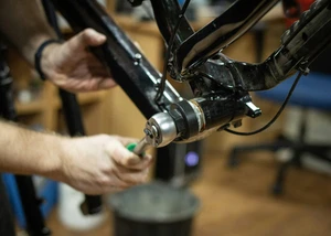 Ein Mechaniker repariert den Rahmen eines schwarzen Fahrrads in einer Werkstatt. Im Fokus steht die präzise Justierung eines Bauteils mithilfe eines Werkzeugs.