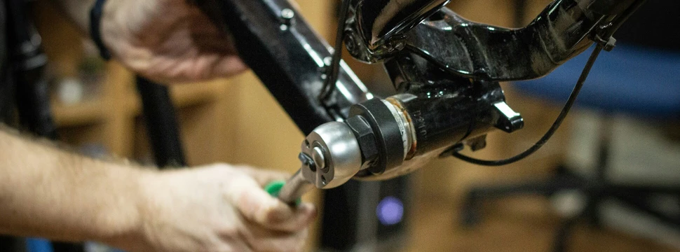 Ein Mechaniker repariert den Rahmen eines schwarzen Fahrrads in einer Werkstatt. Im Fokus steht die präzise Justierung eines Bauteils mithilfe eines Werkzeugs.