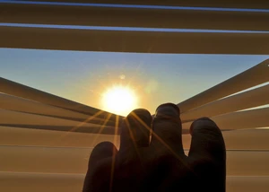 eine Hand öffnet eine Jalousie, durch die die Sonne zu sehen ist