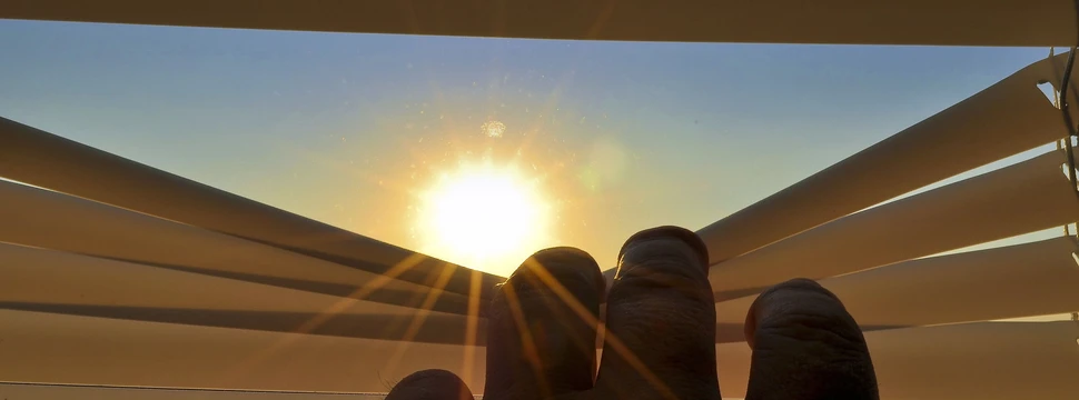 eine Hand öffnet eine Jalousie, durch die die Sonne zu sehen ist