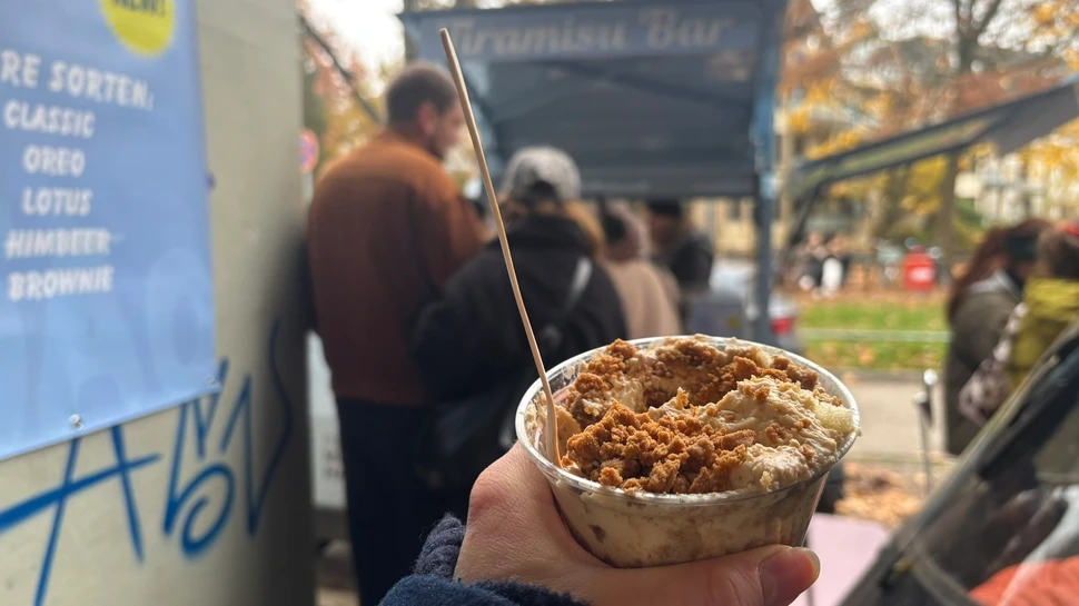 Hand hält einen Becher mit Tiramisu, im Hintergrund undeutlich weitere Menschen