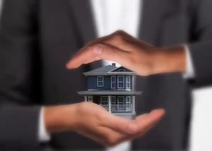Zwei schützende Hände umgeben ein kleines blaues Einfamilienhaus-Modell als Symbol für Immobilienschutz