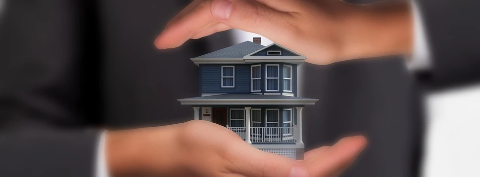 Zwei schützende Hände umgeben ein kleines blaues Einfamilienhaus-Modell als Symbol für Immobilienschutz