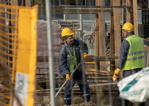 Zwei Bauarbeiter auf einer Baustelle am Arbeiten, durch eine Baustellenabsperrung gesehen