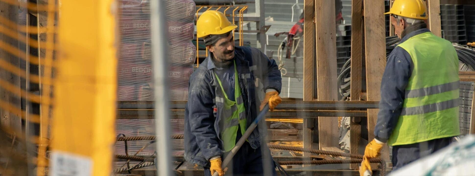 Zwei Bauarbeiter auf einer Baustelle am Arbeiten, durch eine Baustellenabsperrung gesehen