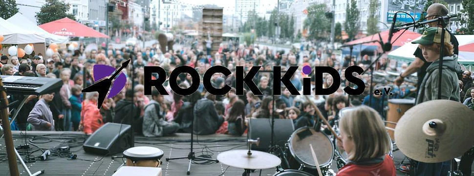 Kinder und Jugendliche musizieren auf einer Bühne im Freien mit Schlagzeug, Percussion und Keyboard vor einem großen Publikum. Im Vordergrund ist ein Kind am Schlagzeug zu sehen. Auf der Bildmitte liegt das Logo „ROCK KIDS e.V.“ mit einer stilisierten Gitarre. Im Hintergrund stehen viele Menschen und bunte Pavillons in einer städtischen Umgebung.