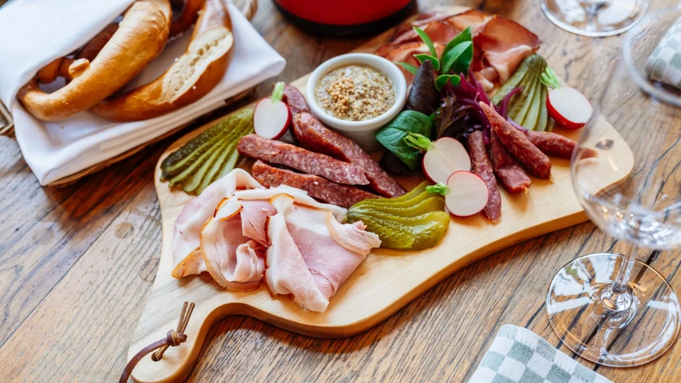 Eine appetitlich angerichtete Aufschnitt-Platte mit Wurst und Schinken auf rustikalem Holztisch. Aufschnitt-Platte mit Wurst, Schinken, Gurken und Kräutern auf Holzbrett, dazu Brezeln und Weingläser.