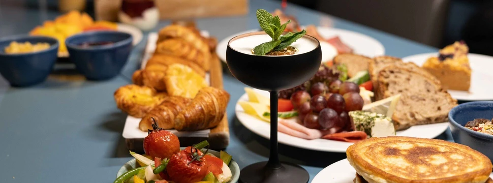 Reichhaltig gedeckter Brunch-Tisch mit Pancakes, Croissants, Käse-Wurst-Platte und Cocktail im nordischen Stil