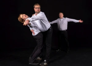 Drei Improvisationsschauspieler in weißen Hemden und dunklen Hosen bei einer dynamischen Theaterszene.