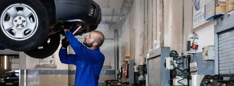 Ein Mitarbeiter leuchtet in einer Autowerkstatt unter ein Auto, welches auf einer Hebebühne liegt