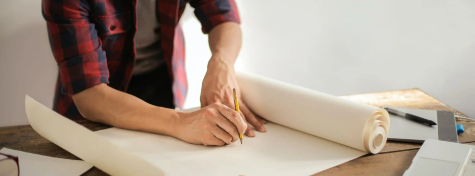 Person in kariertem Hemd zeichnet mit Bleistift auf Tapetenrolle am Holztisch mit Arbeitsutensilien