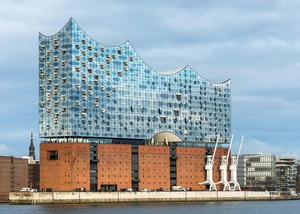 Die Elbphilharmonie in Hamburg mit ihrer markanten gläsernen Fassade, die sich im Tageslicht spiegelt. Im Vordergrund sind der Hafen und drei weiße Hafenkräne zu sehen.