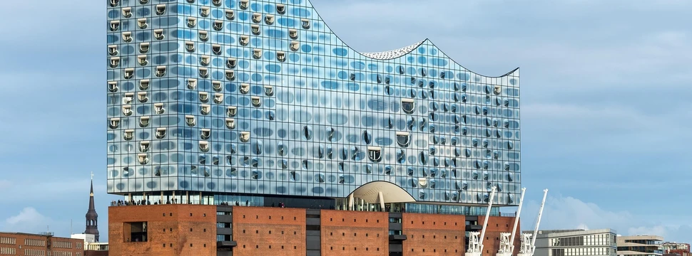 Die Elbphilharmonie in Hamburg mit ihrer markanten gläsernen Fassade, die sich im Tageslicht spiegelt. Im Vordergrund sind der Hafen und drei weiße Hafenkräne zu sehen.