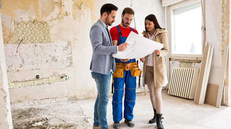Ein typisches Beratungsgespräch des BeBM vor Ort 3 erwachsene Menschen mit einem Bauplan auf einer Baustelle eines Hauses
