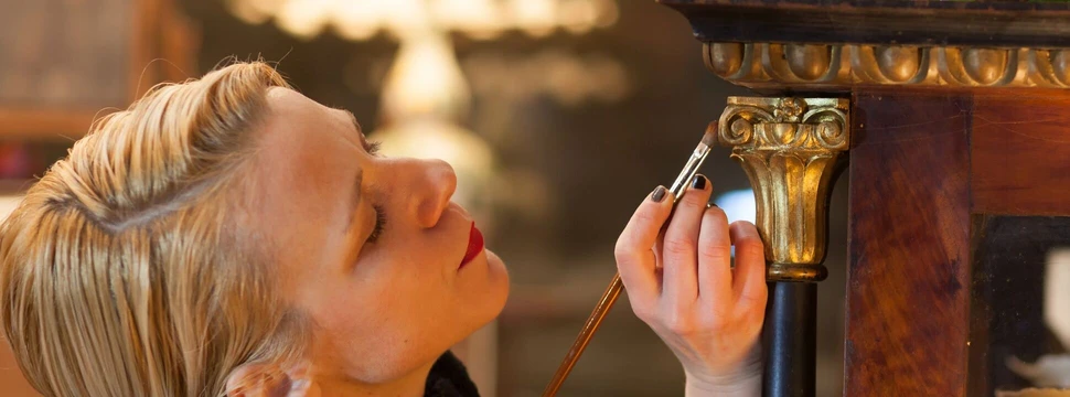 Möbel-Restauratorin malt mit einem Pinsel Goldfarbe auf einen Schrank