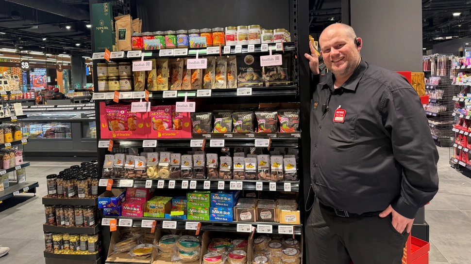 Regal im Supermarkt, daneben ein Mitarbeiter in schwarzer Kleidung