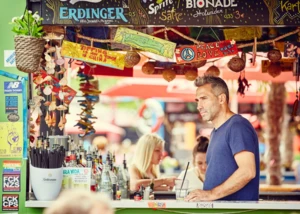Mann in blauem Shirt an bunter Container-Bar mit Getränkeflaschen und Peace-Love-Schildern am Spielbudenplatz.