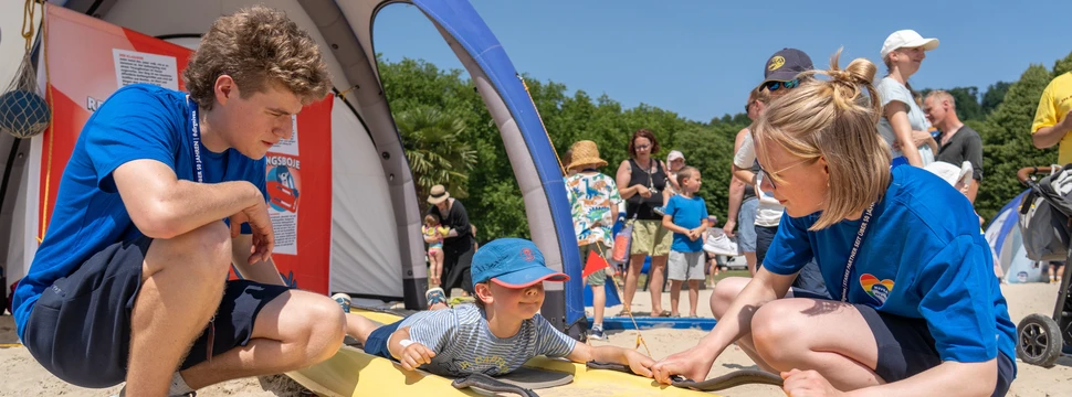 Ein Kind liegt auf einem gelben DLRG-Rettungsboard im Sand und wird von zwei jungen Helfern in blauen T-Shirts betreut. Im Hintergrund stehen weitere Besucher und Kinder an einem Informationszelt.
