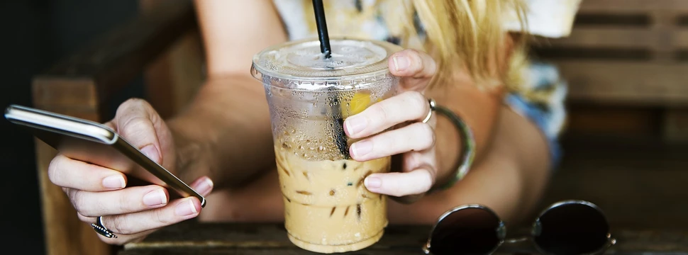 Frau hält in der einen Hand ein Handy und in der anderen Hand einen Iced Coffee. Daneben liegt eine Sonnenbrille