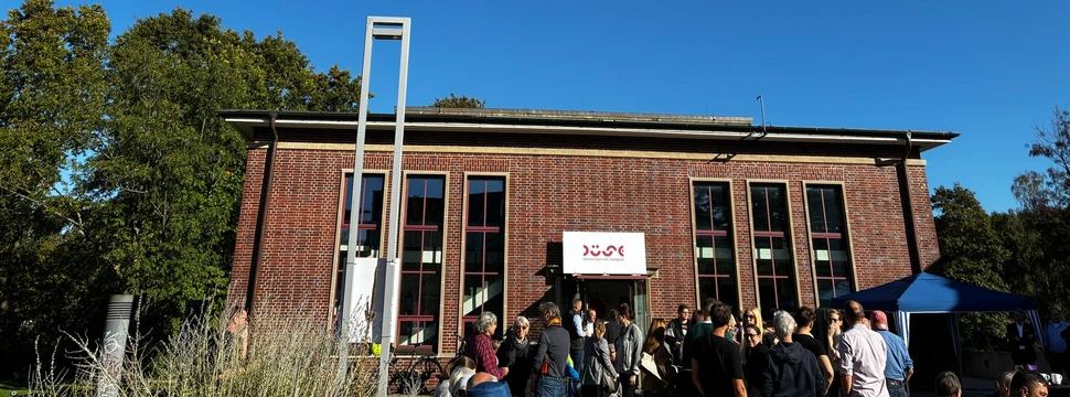 Eine Gruppe von Menschen vor einem rotgeklinkertem Haus mit Sonne und blauem Himmel