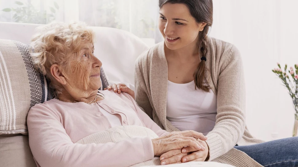 Pflegedienste helfen bei der häuslichen Pflege. Jüngere Frau hält die Hand einer Seniorin auf dem Sofa.