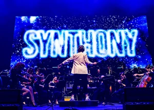 Ein Orchester spielt auf der Bühne, im Hintergrund leuchtet das Wort "Synthony"