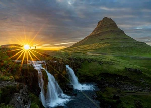 Eine Landschaftaufnahme von einem Berg, davor ist ein Wasserfall, und die Sonne geht gerade am Horizont unter