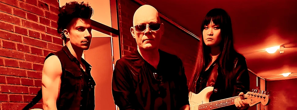 Die Band The Sisters of Mercy posiert in einem rot beleuchteten Flur mit Gitarren.