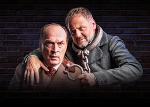 Eine intensive Theaterszene mit Samuel Finzi und Herbert Knaup vor industrieller Backsteinwand. Zwei Männer in einer theatralischen Pose mit Handschellen vor einer dunklen Backsteinwand in dramatischer Beleuchtung.