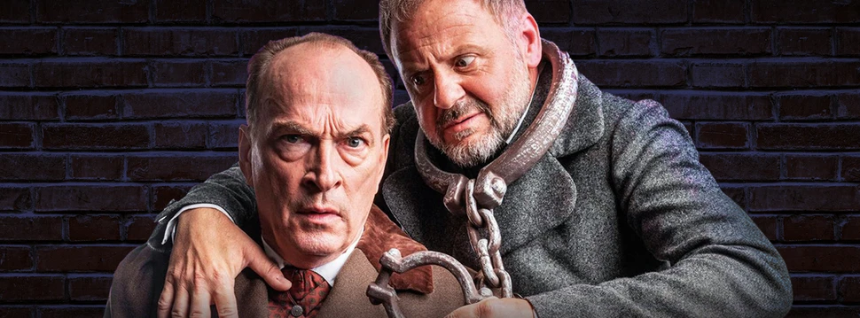 Eine intensive Theaterszene mit Samuel Finzi und Herbert Knaup vor industrieller Backsteinwand. Zwei Männer in einer theatralischen Pose mit Handschellen vor einer dunklen Backsteinwand in dramatischer Beleuchtung.