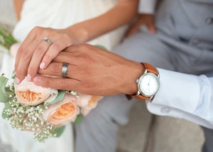 Das Bild zeigt die Hände eines frisch verheirateten Paares, das zärtlich die Ringe präsentiert. Im Hintergrund ist ein Brautstrauß mit zarten Rosen zu sehen.