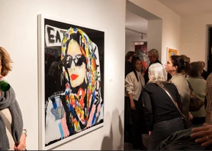 Besuchende in einer Galerie betrachten ein Bild von einer Frau mit Sonnenbrille und buntem Kopftuch