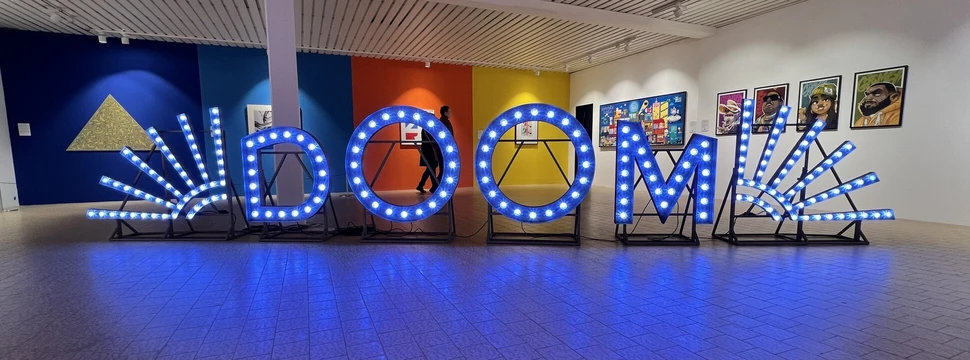 Das Wort "Doom" als Leuchtreklame steht in einem Raum mit anderen Kunstwerken an der Wand