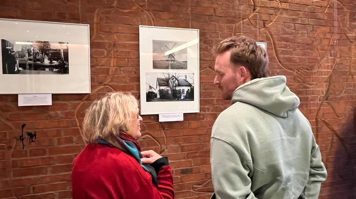 Galerie-Bild 1: Zwei Erwachsene im Gespräch vor der Wand mit den verschiedenen Fotos