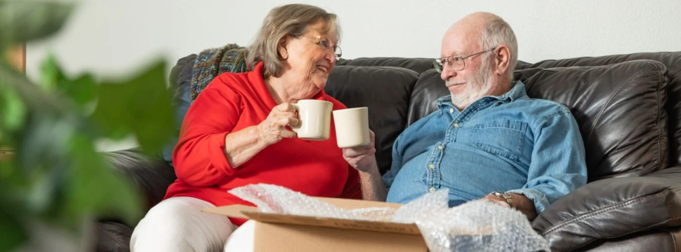 Ein älteres Paar sitzt mit Kaffeebechern auf einer Couch und stößt an, davor stehen Umzugskartons.