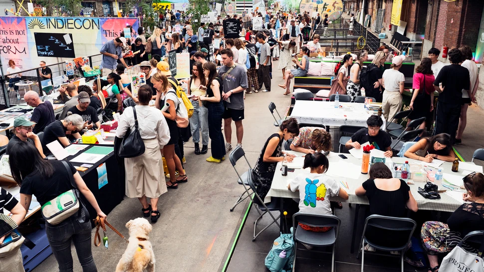 Indiecon Stände Eine große Halle mit industriellem Charme voller Stände und Tische, an denen zahlreiche Menschen Bücher, Magazine, Kunstwerke und Druckprodukte präsentieren und betrachten. Viele Besucher schlendern durch die Reihen oder unterhalten sich mit den Ausstellenden. Im Hintergrund ist ein großes farbiges Banner mit der Aufschrift „INDIECON“ zu sehen. Oberhalb verlaufen Stahlträger und Lichtinstallationen unter dem Dach. Die Atmosphäre wirkt lebendig und kreativ, wie auf einer Messe oder einem Festival für unabhängige Verlage und Künstler.