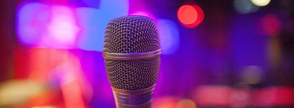 Ein Mikrofon im Fokus vor unscharfen, bunten Bühnenlichtern in violett, rot und orange Tönen.