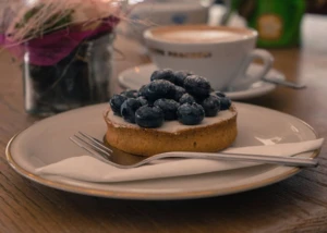 Blaubeer-Törtchen auf weißem Teller mit Goldrand, Kuchengabel und Serviette, im Hintergrund eine Cappuccino-Tasse und ein Vasen-Arrangement mit Trockenblumen.