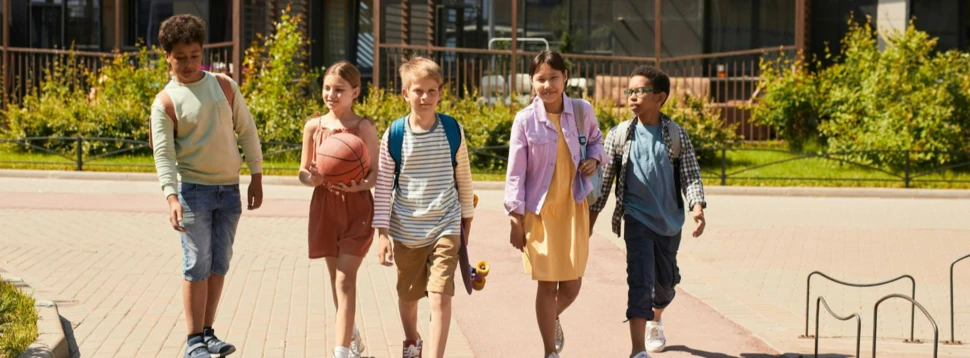 Schulkinder mit Skateboard und Basketball auf dem modernen Schulhof unterwegs. Fünf diverse Schulkinder gehen über sonnigen Schulhof, einer auf Skateboard, eine mit Basketball.