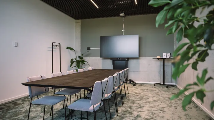 Galerie-Bild 1: Moderner Konferenzraum mit Holztisch, grauen Stühlen, Wandmonitor und Grünpflanzen in minimalistischem Design.