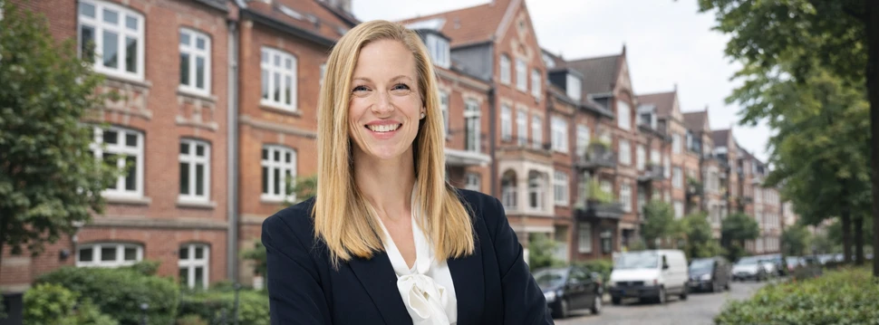 Eine Frau in einem Anzug und weißem Hemd vor einer Reihe aus Häusern, die aus roten Backsteinen bestehen
